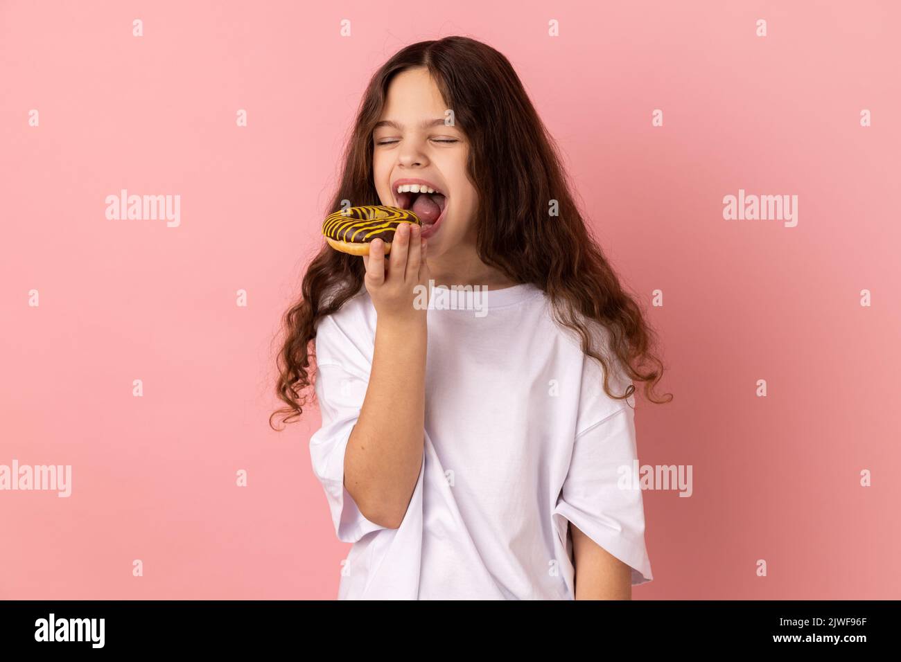 Sugar addiction. Portrait of satisfied little girl wearing white T-shirt biting delicious donut, looking with desire to eat sweet dessert. Indoor studio shot isolated on pink background. Stock Photo