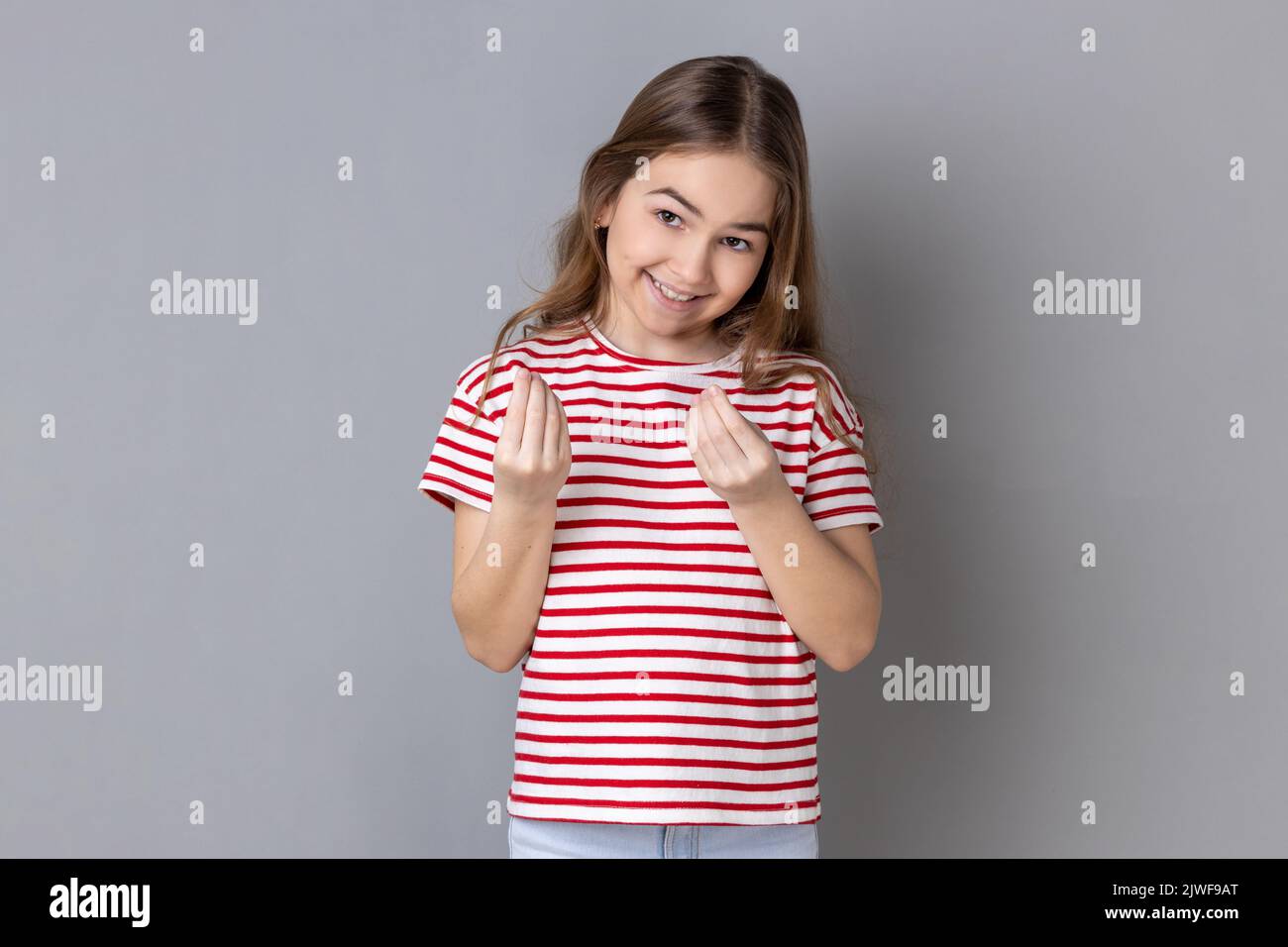 Portrait of smiling little girl wearing striped T-shirt makes money gesture, rubs fingers, looking at camera with glad expression. Indoor studio shot isolated on gray background. Stock Photo