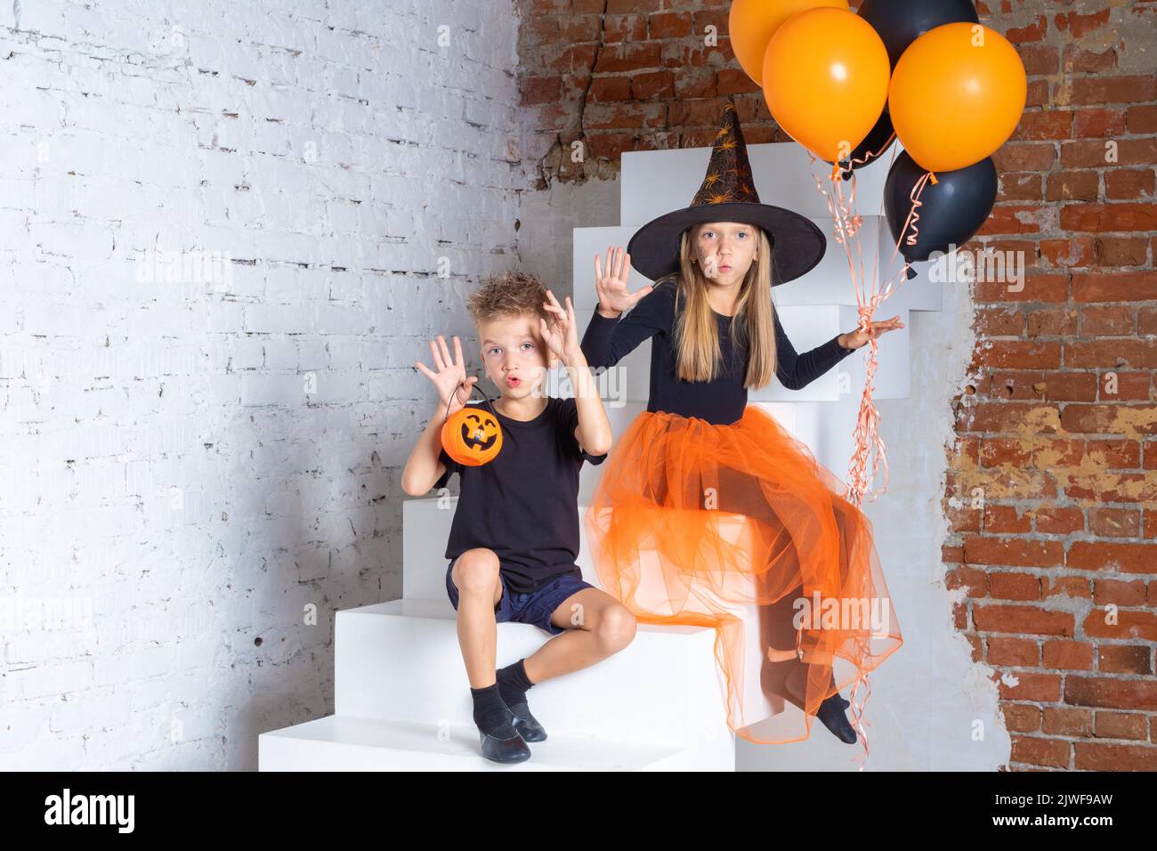 Kids on Halloween. A little girl and a boy in witch and sorcerer costumes with hats holding orange and black balls and making frightening gestures wit Stock Photo