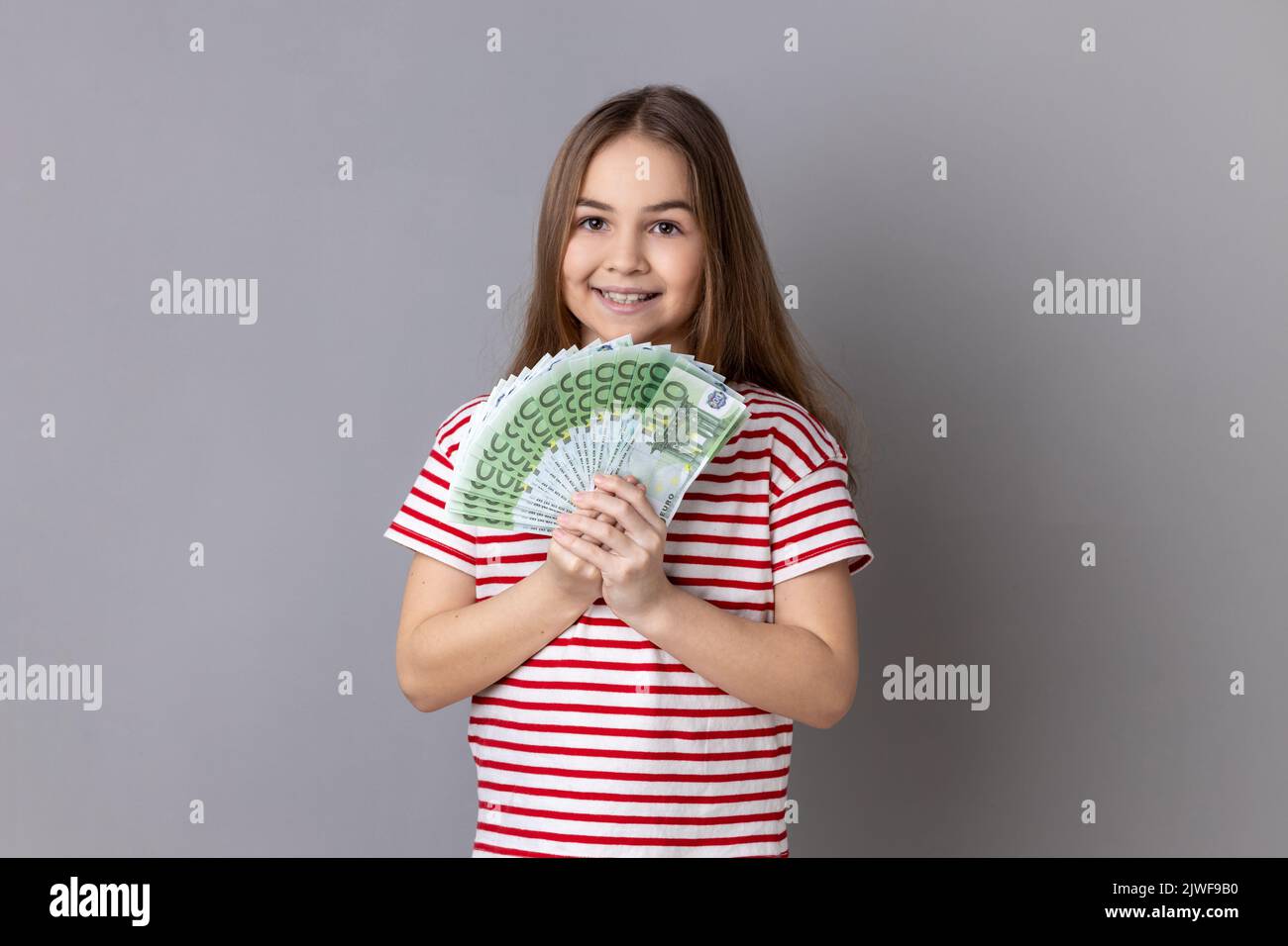 Portrait of satisfied little girl wearing striped T-shirt holding at fan of euro banknotes, showing big money and looking at camera. Indoor studio shot isolated on gray background. Stock Photo