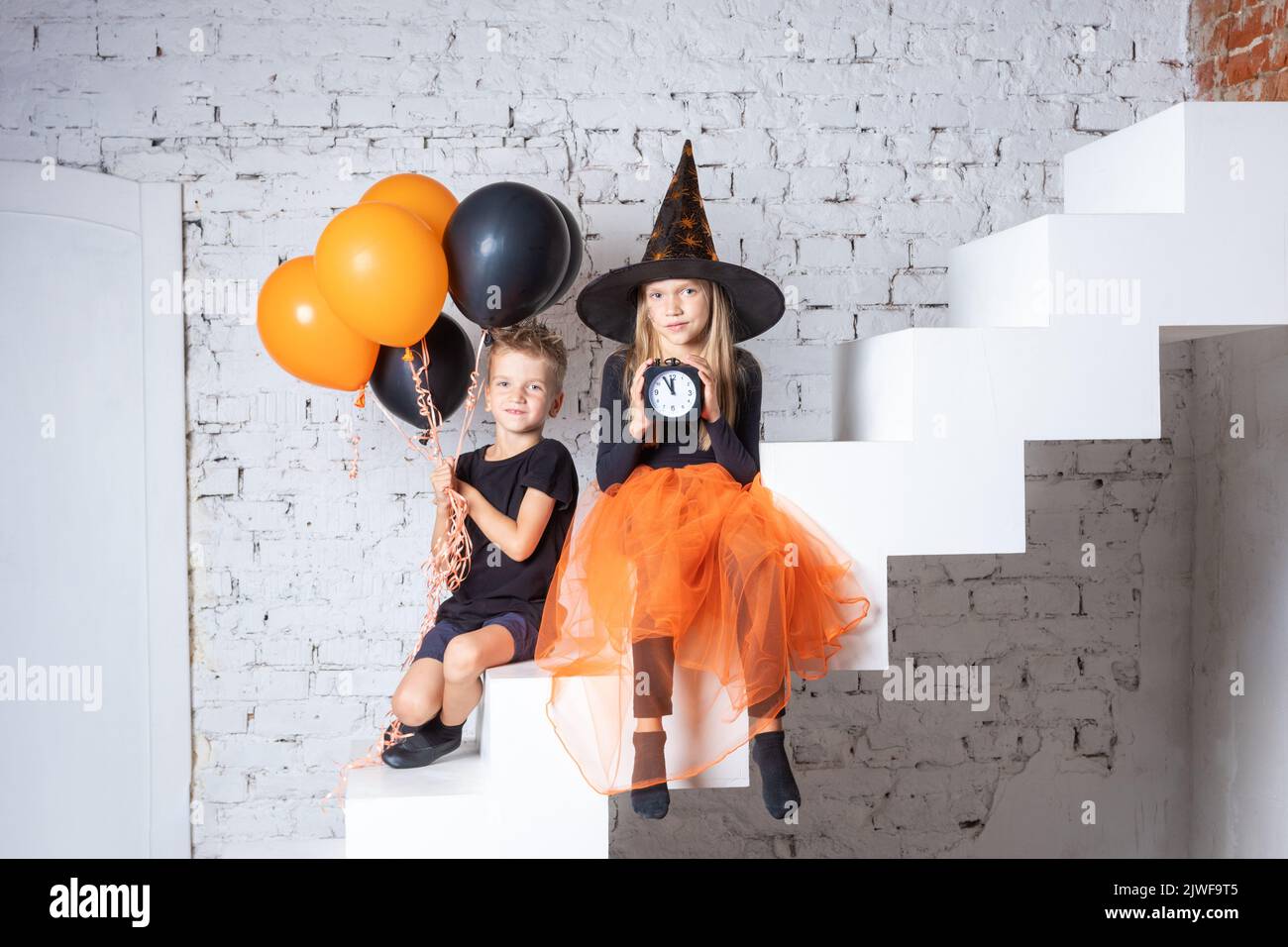 Halloween time. Children in creepy witch costumes and hats are holding a black alarm clock and black and orange balloons while sitting on the stairs. Stock Photo