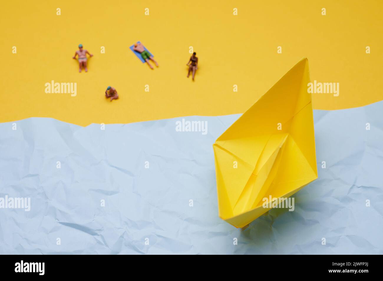 yellow paper boat on a blue background and miniature people sit on a yellow paper which symbolizes the beach Stock Photo