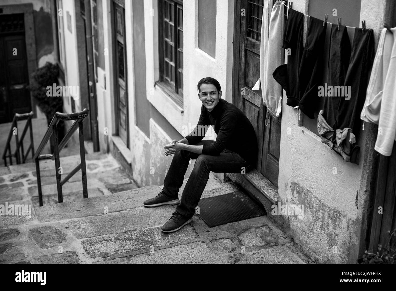 Handsome young man sitting on the steps of a narrow street in an old downtown European city. Black and white photograph. Stock Photo