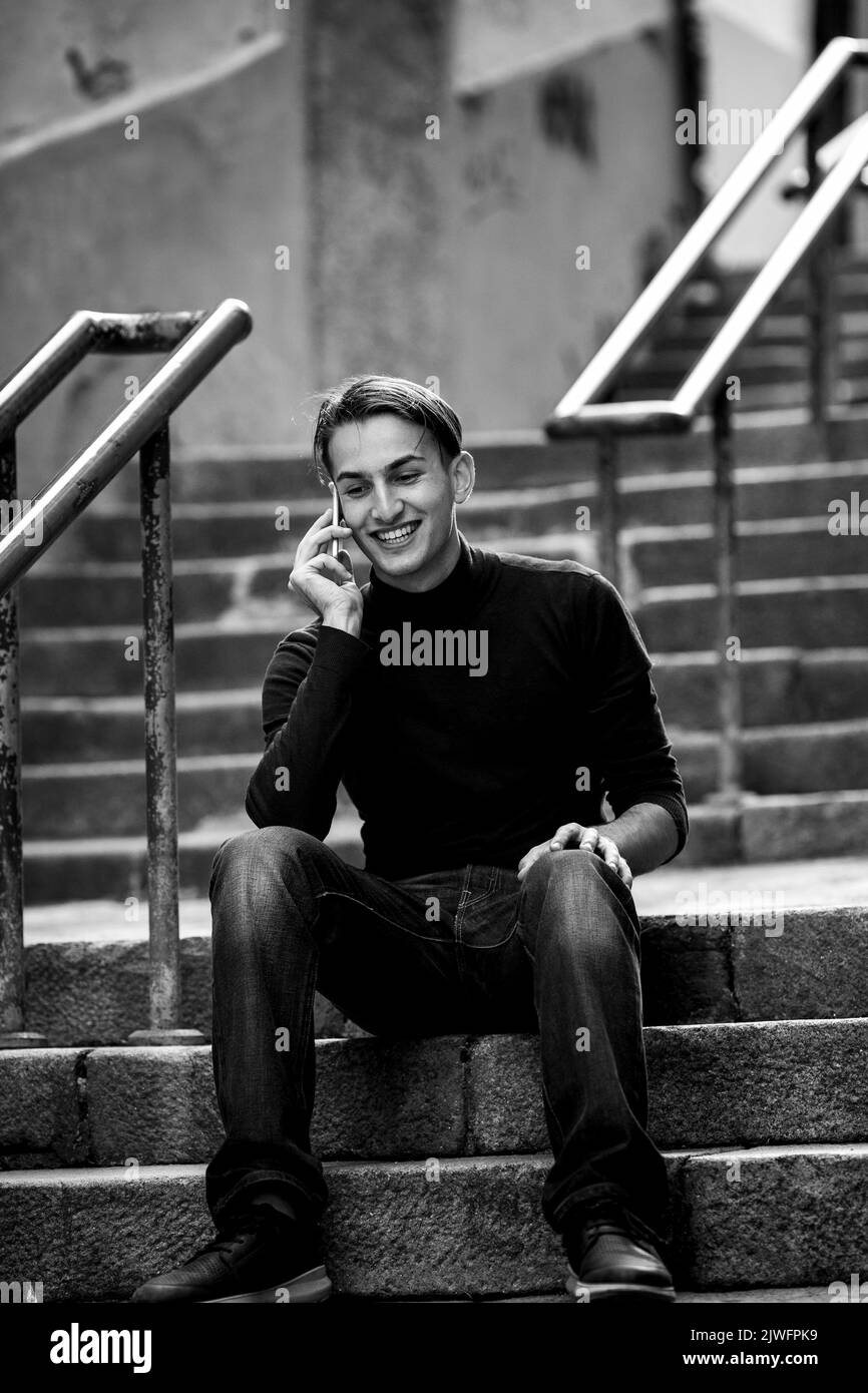 A handsome young man sitting on the steps on a narrow street talking on his cell phone. Black and white photo. Stock Photo