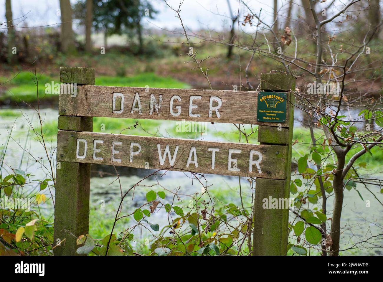 A wooden sign warns people of the danger of deep water near a small pond in a woodland area in North London in England UK Stock Photo