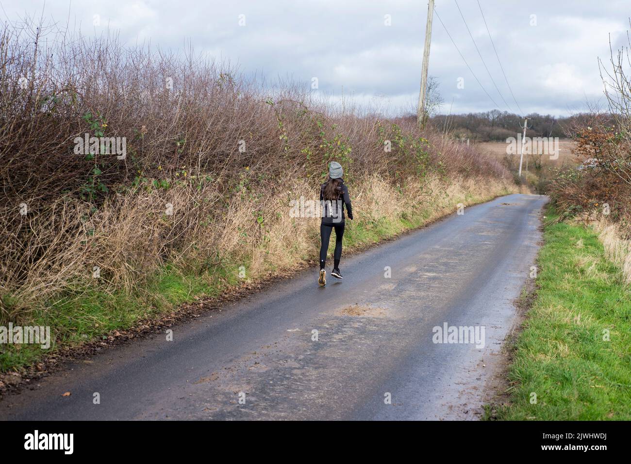 A female runs along a country lane by a tall hedgerow on a tarmac road in the countryside of North London in England Stock Photo