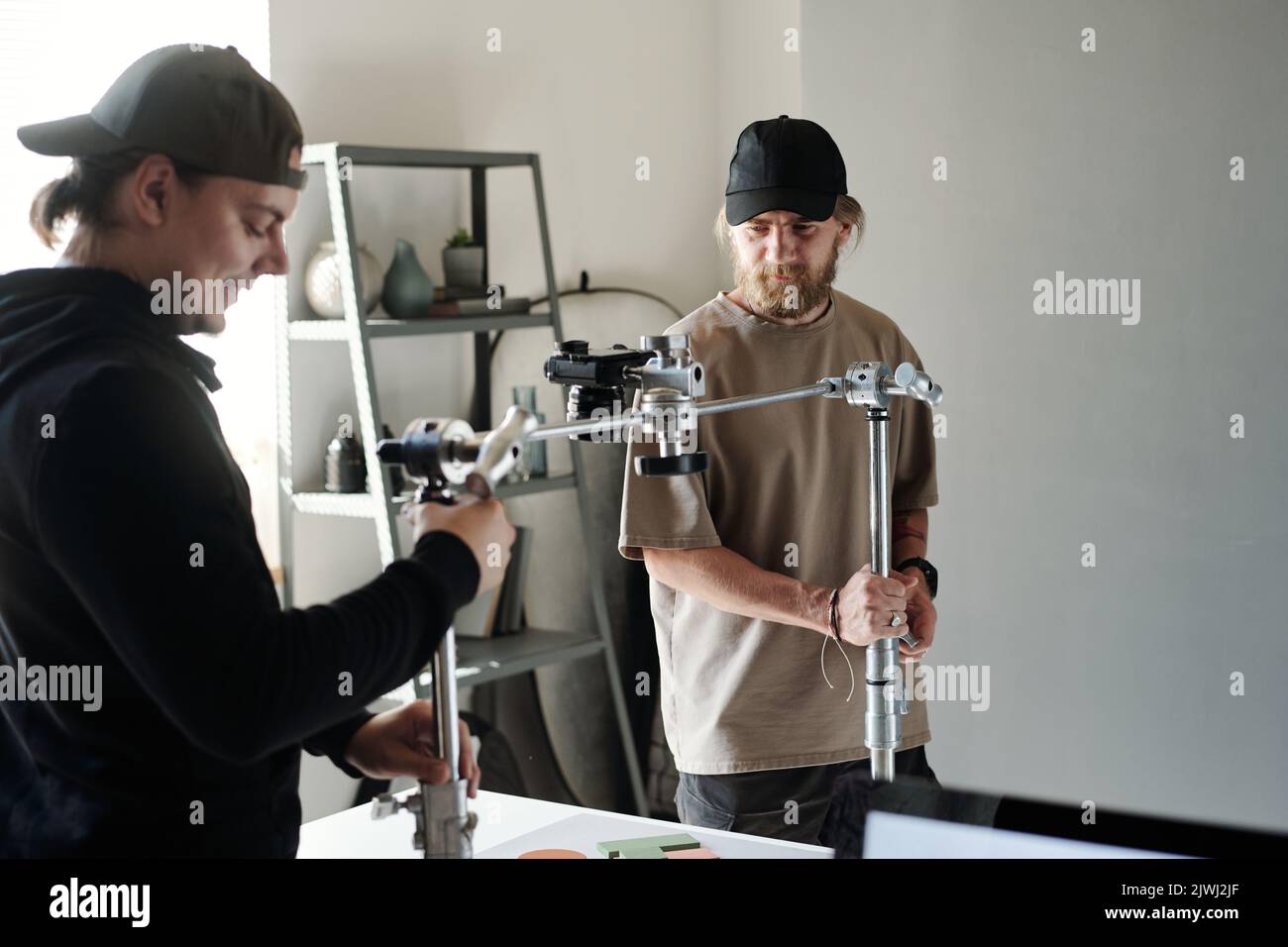 Young assistant helping photographer or videographer adjust steadicam and metallic slider over table to shoot new commercial Stock Photo
