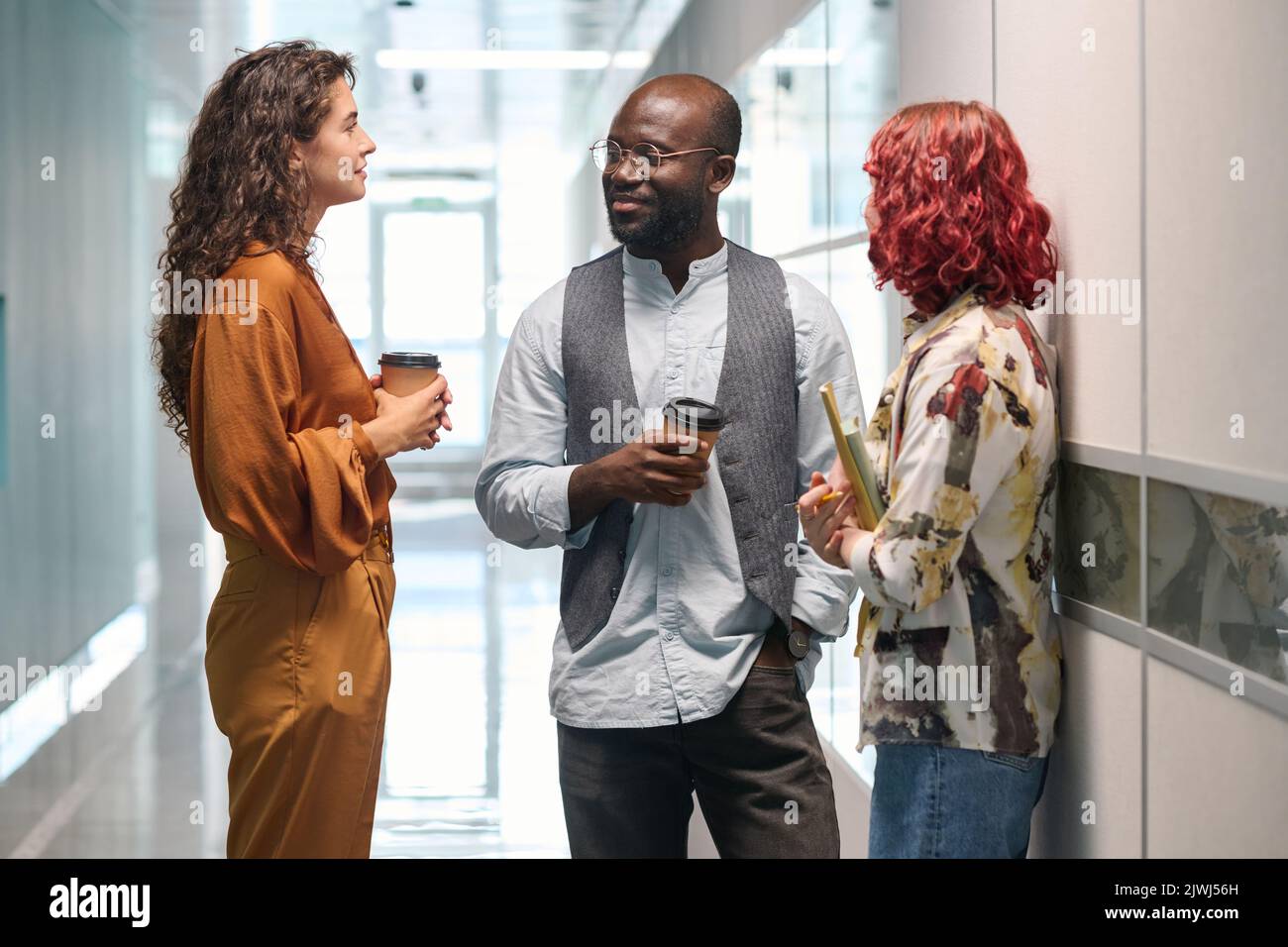 Three young successful office managers in casualwear having chat while standing between two walls in corridor of modern building Stock Photo