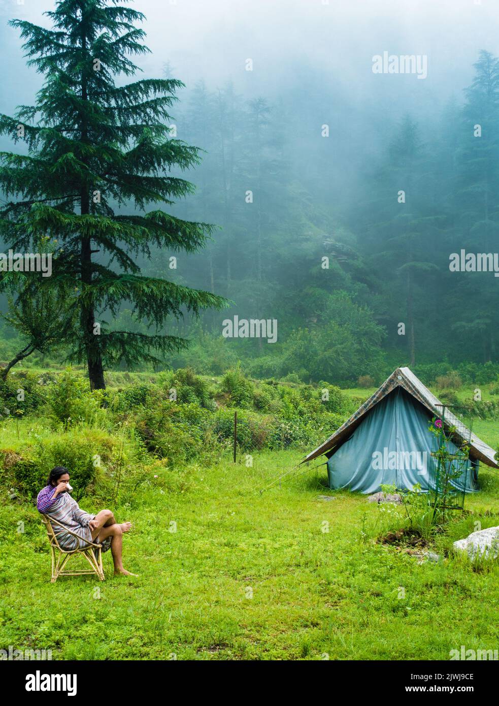 July 7th 2021. A man sipping on a hot tea cup in the meadows with a camp amid deodar tree forest and mountains in the background. Uttarakhand India. Stock Photo