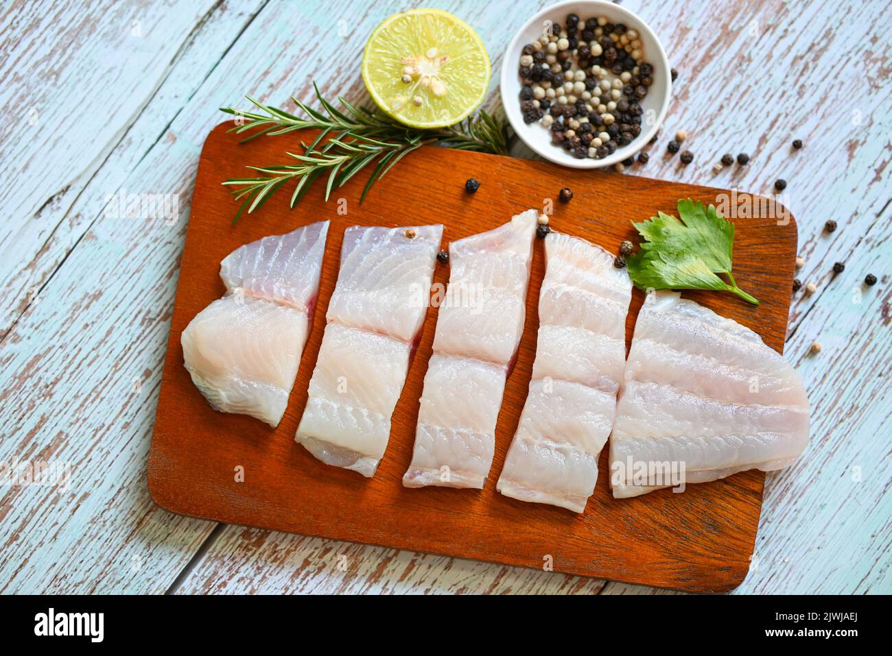 fresh raw pangasius fish fillet with herb and spices black pepper lemon lime and rosemary, fish fillet on wooden board with ingredients for cooking, m Stock Photo