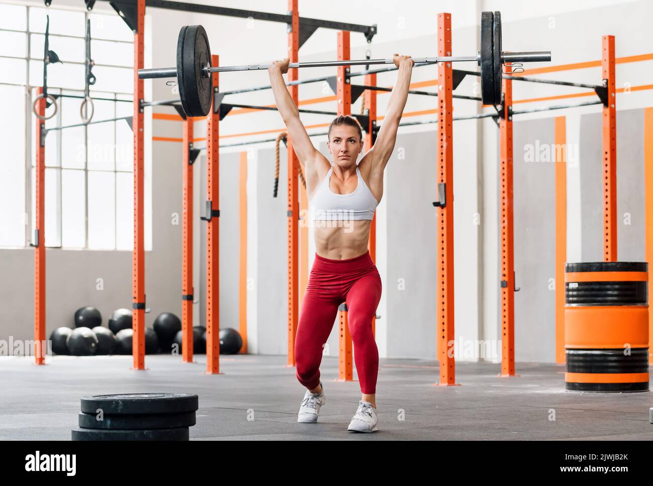 Full body strained sportswoman lifting heavy barbell over head and lunging while doing split jerk exercise during intense weightlifting training in gy Stock Photo