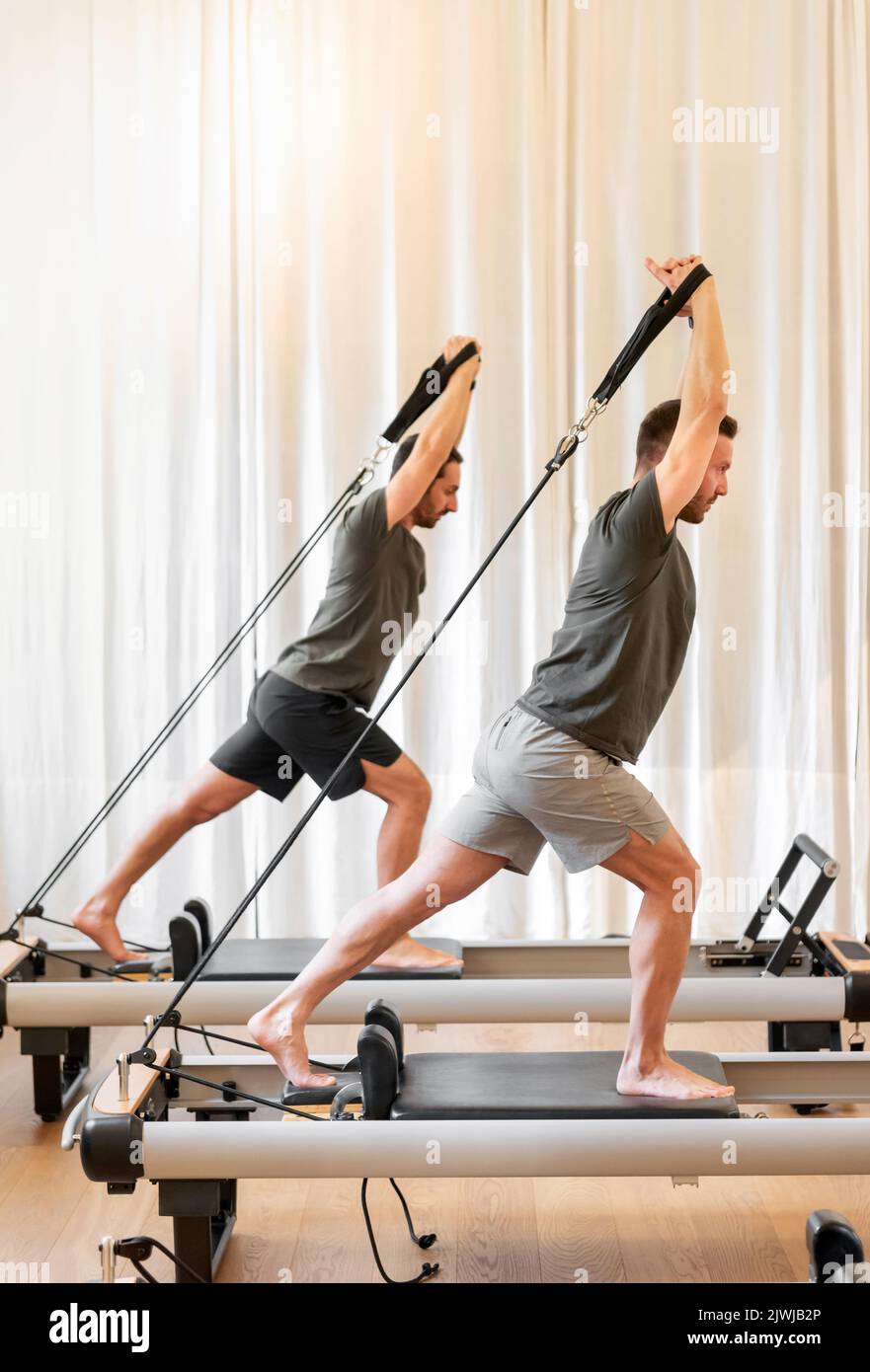 Full body side view of sportive men doing pilates saute exercise on reformer beds during training together in light gym Stock Photo