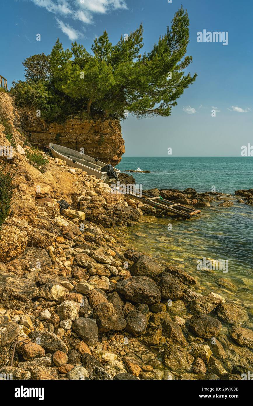 The stony coast of the Chianca Masitto beach. A small jetty with a boat pulled out of the water topped by a pine tree. Monte Sant'Angelo, Puglia Stock Photo