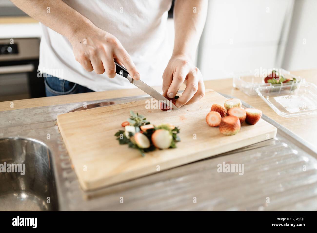 A man wearing a white t-shirt chopping up strawberries in a kitchen on a wooden board with a knife Stock Photo
