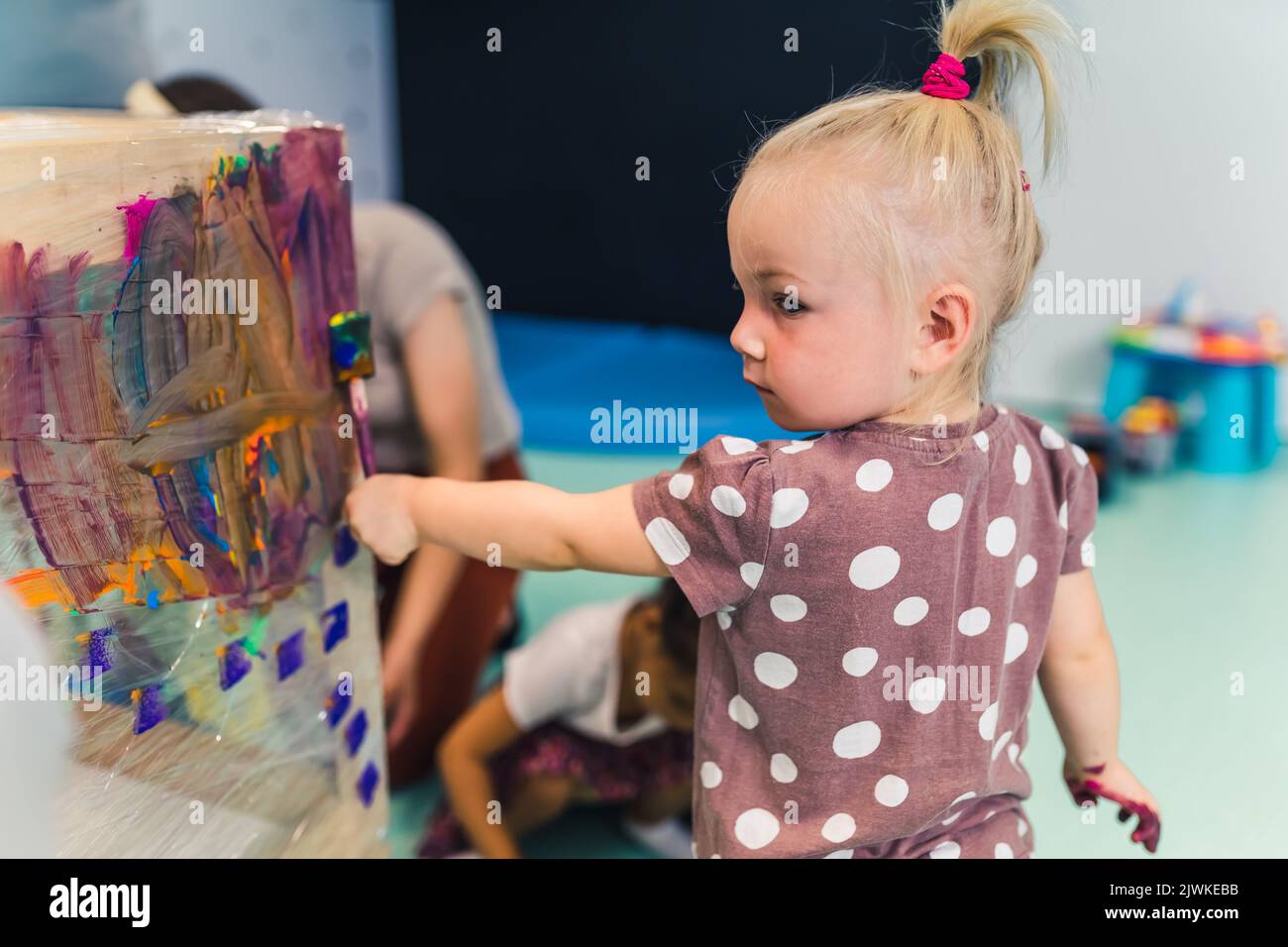Cling film painting. Little caucasian girl toddler painting with her arms on a cling film wrapped all the way round the wooden shelf unit. Creative activity for kids development at the nursery school. High quality photo Stock Photo
