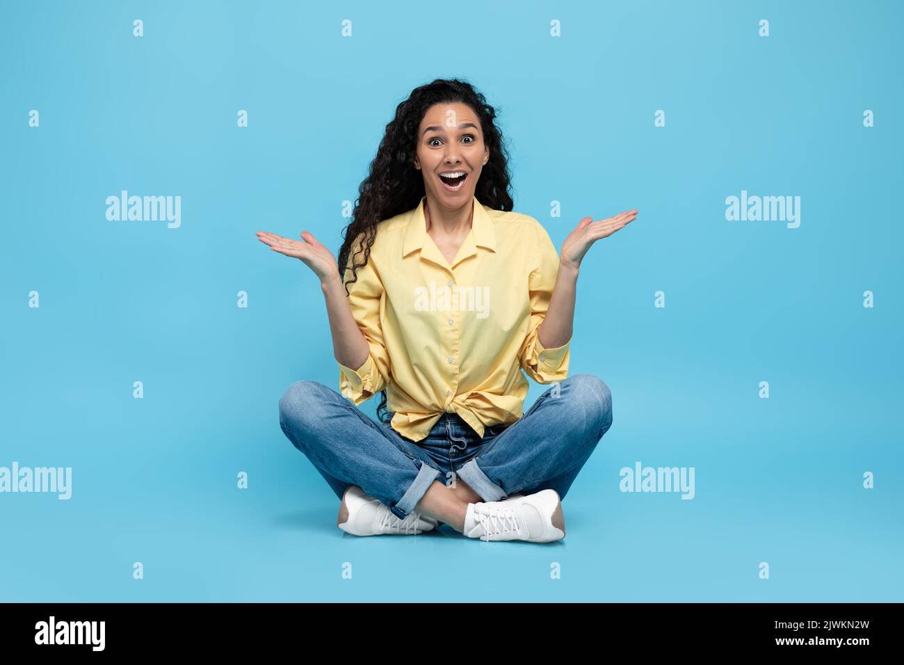 Puzzled excited young woman shrugging shoulders and smiling at camera on blue studio background, full length Stock Photo