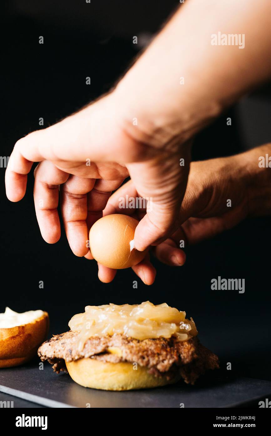 Human hand opening a boiled egg and pouring it on a Doble smashburger with coocked onion over a black background Stock Photo