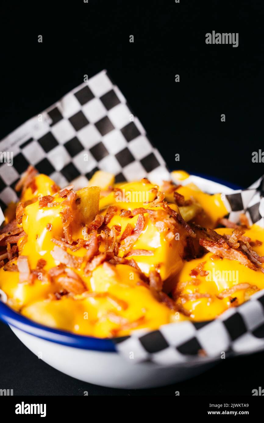 Fried potatoes with melted cheese and topped with pieces of bacon on a white plate over a black background Stock Photo