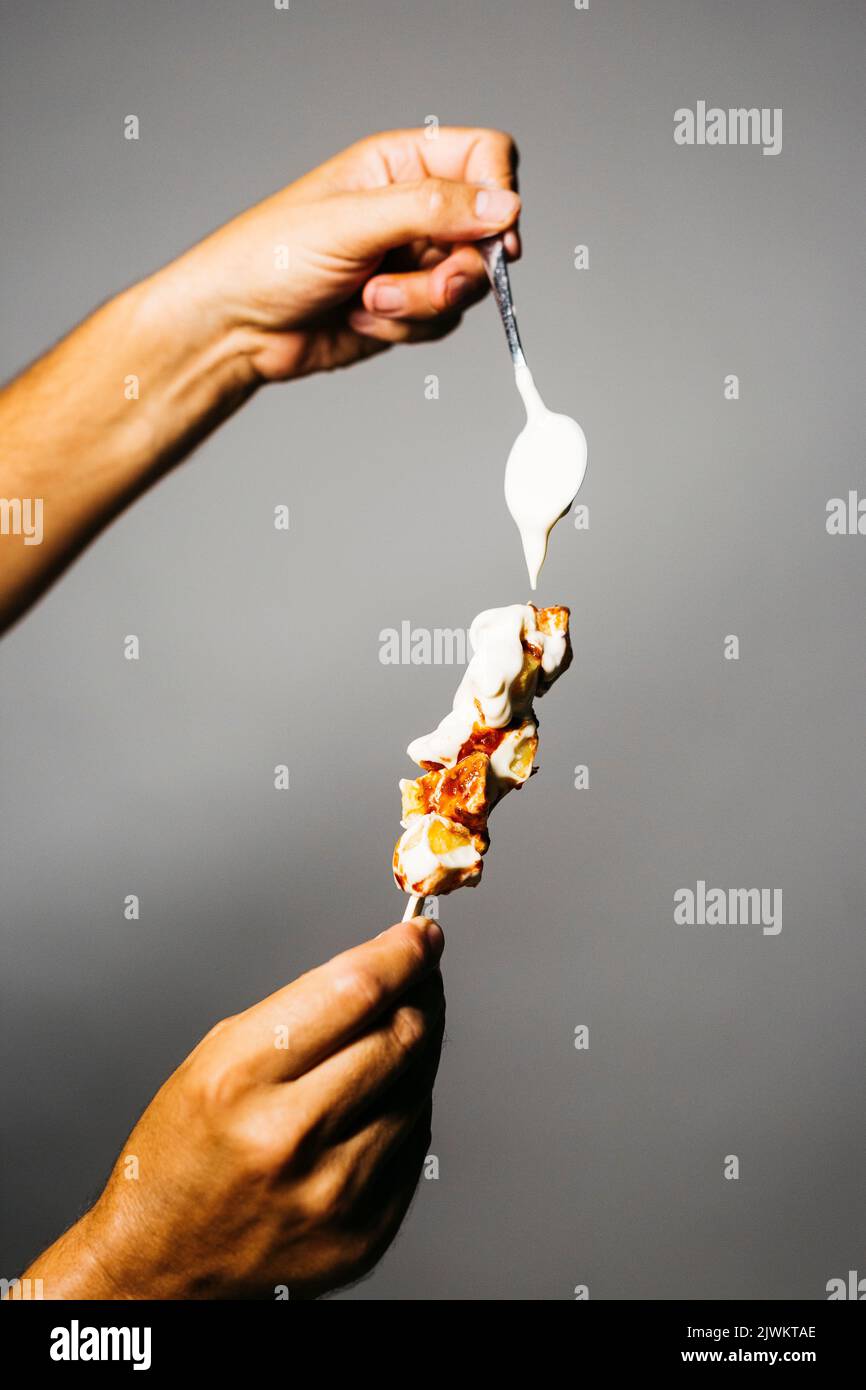 Human hand holding a brochette of fried potatoes with mayonnaise and tomato spacy tomato sauce over a grey background Stock Photo