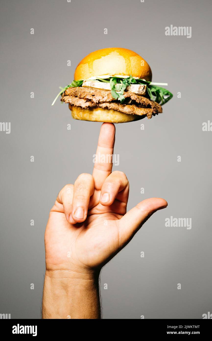 Human hand holding, with his middle finger, a Doble burger with old dry cheese and lettuce over a grey background Stock Photo