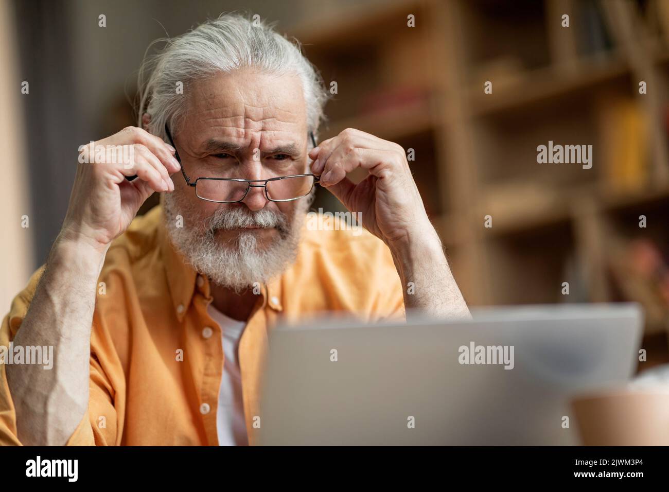 Concentrated old man using notebook at home, touching his eyeglasses Stock Photo