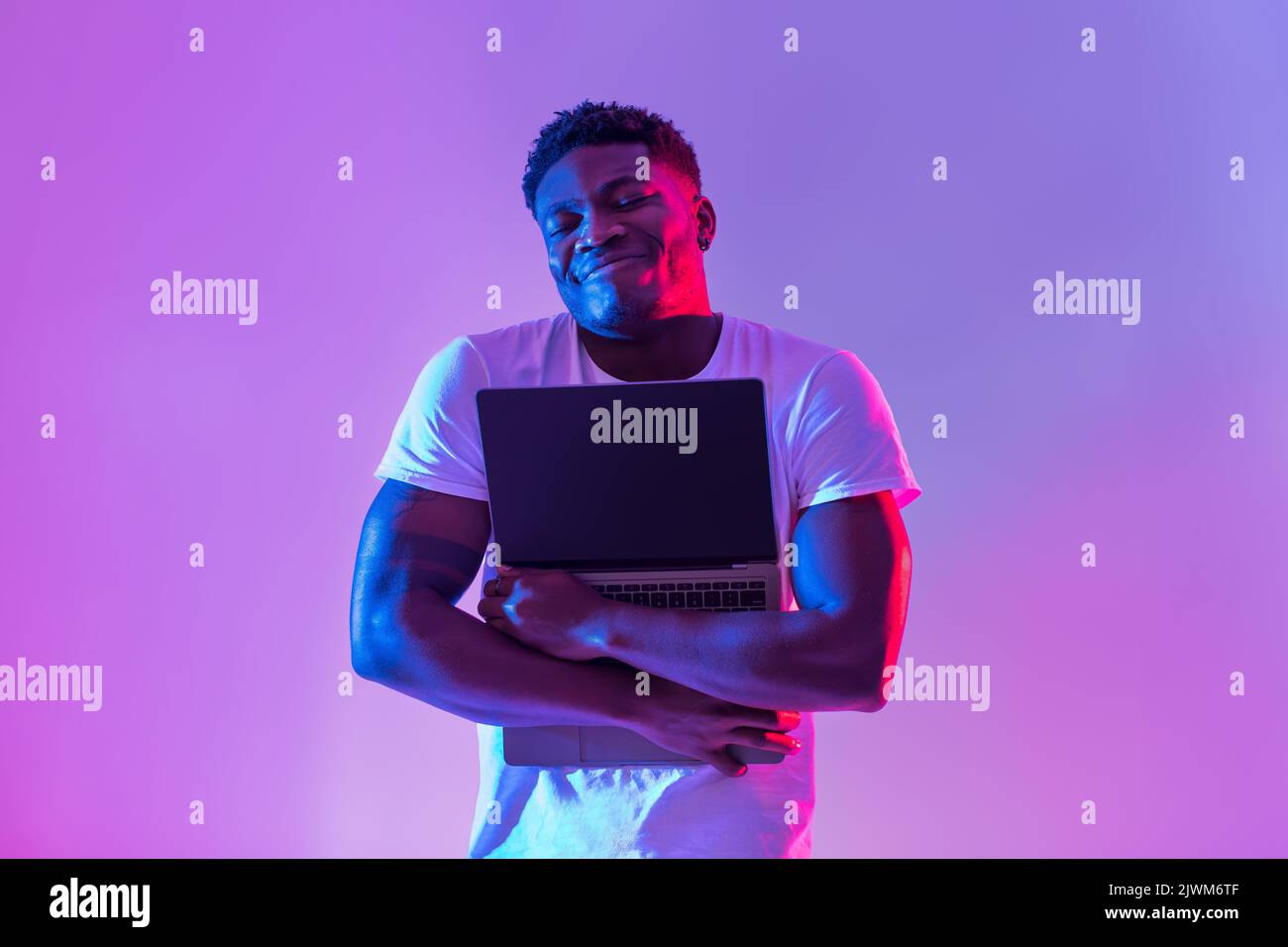 Cheerful young black man hugging laptop with empty screen in neon light ...