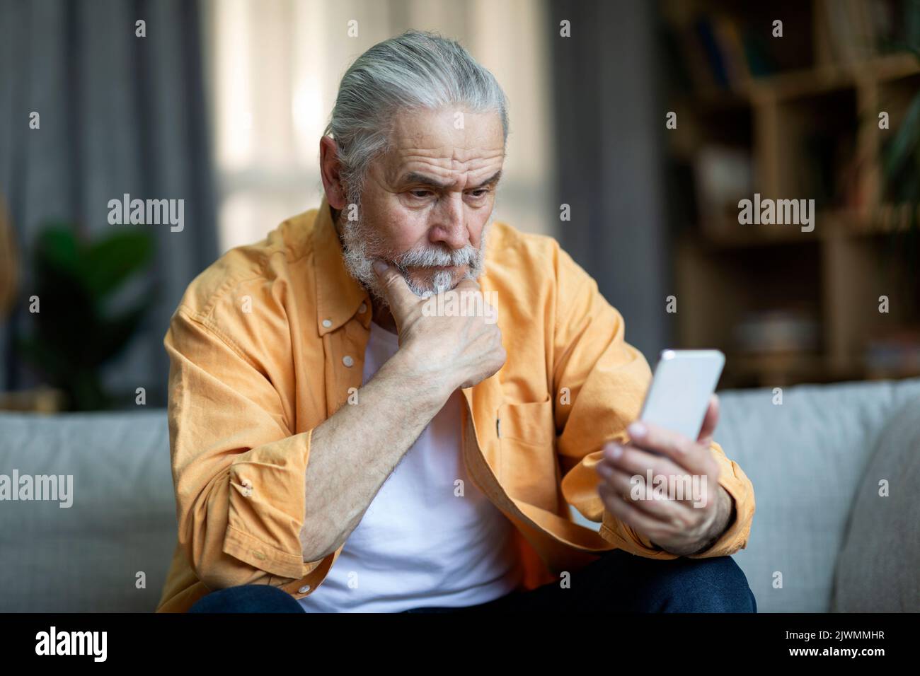 Pensive elderly man sitting on couch, using smartphone Stock Photo
