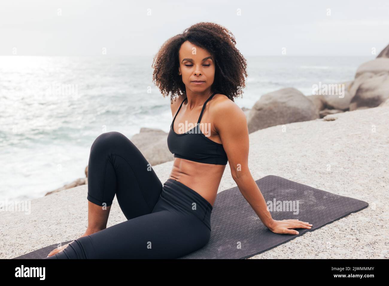 Fit woman with closed eyes sitting on mat by ocean relaxing after yoga training Stock Photo