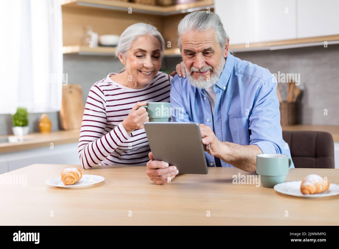 Happy Senior Spouses Using Digital Tablet Together While Having Breakfast In Kitchen Stock Photo