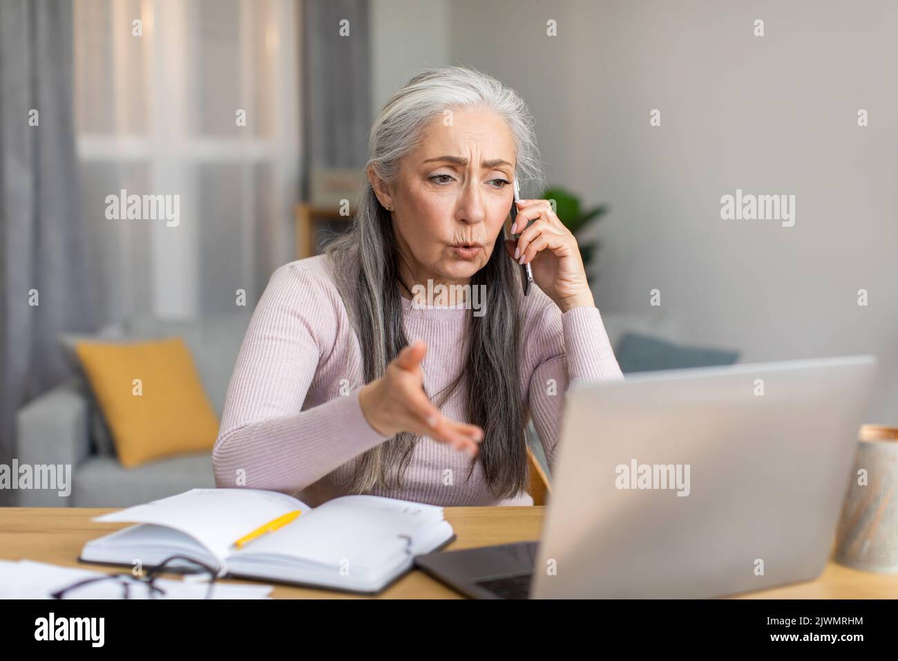 Sad serious caucasian elderly gray-haired woman speaks by phone and looks at laptop with client Stock Photo