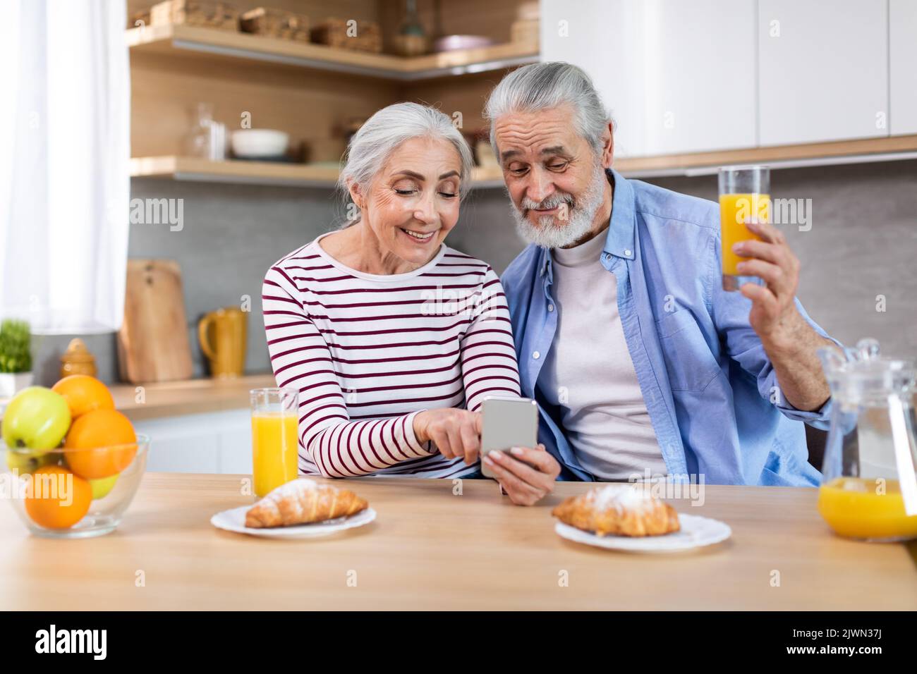 Happy Senior Couple Using Smartphone In Kitchen While Having Breakfast Together Stock Photo