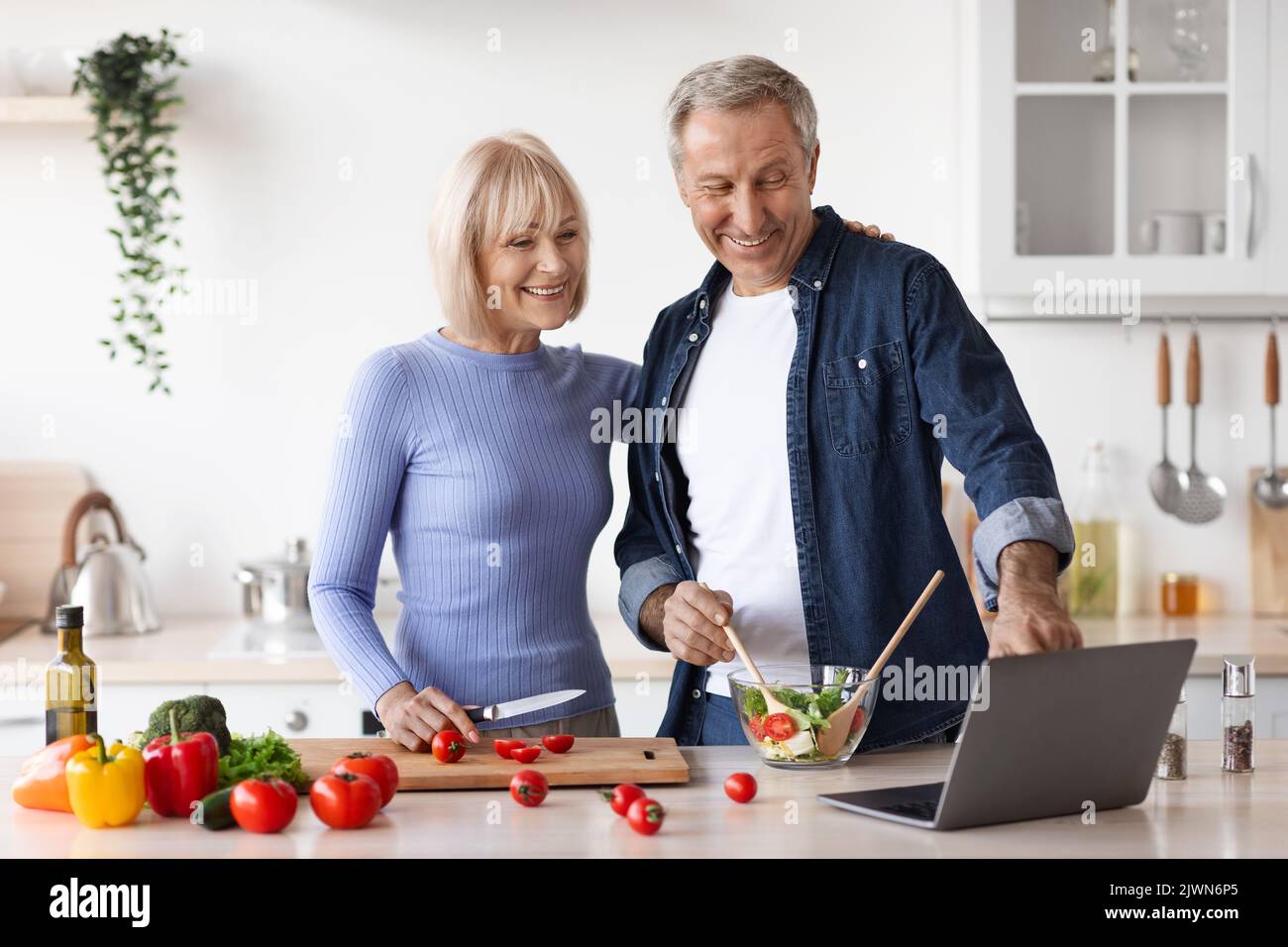 Loving senior spouses cooking together at home, using laptop Stock Photo