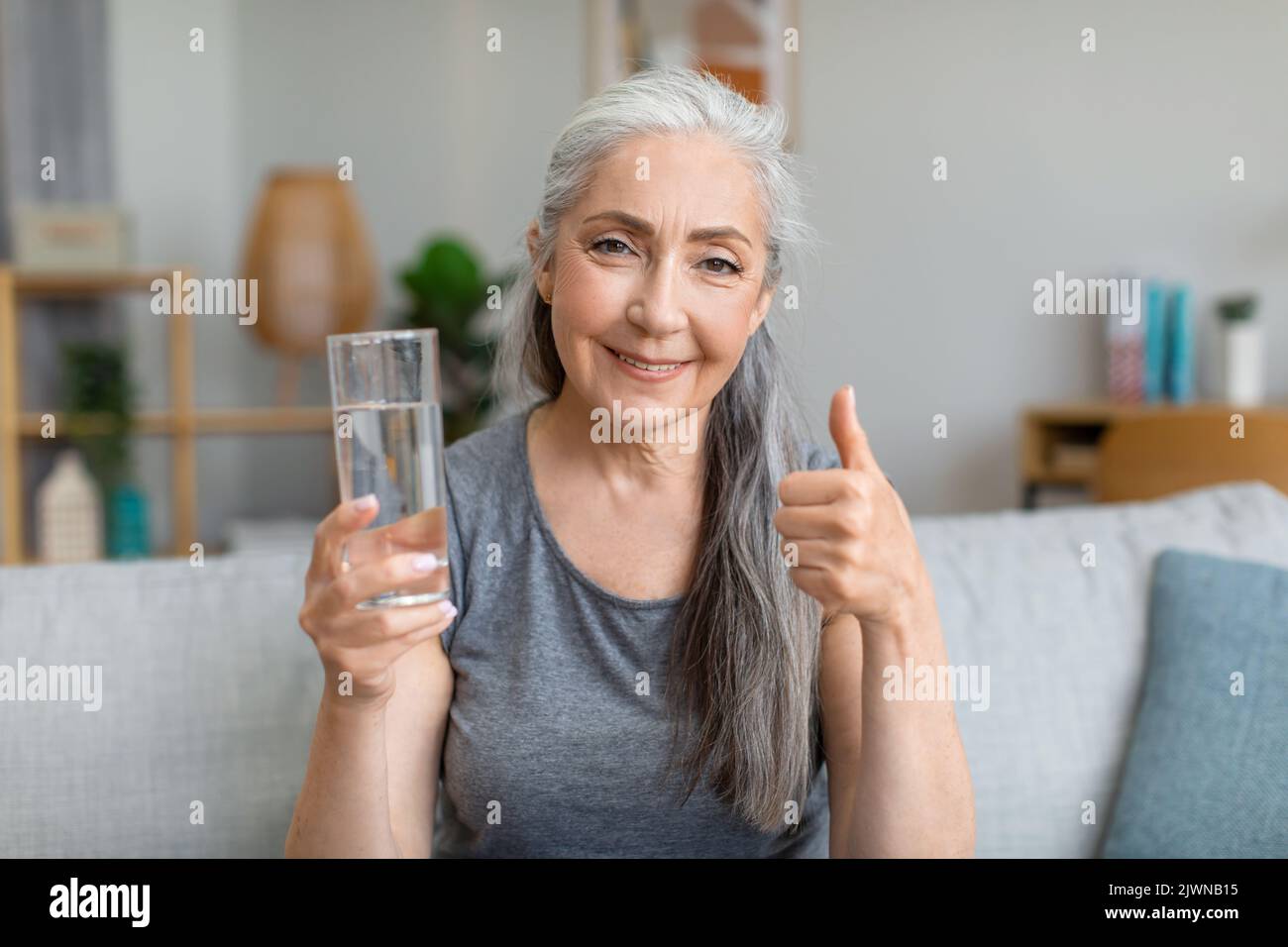Happy pretty caucasian senior gray-haired female holds glass of water and shows thumb up Stock Photo