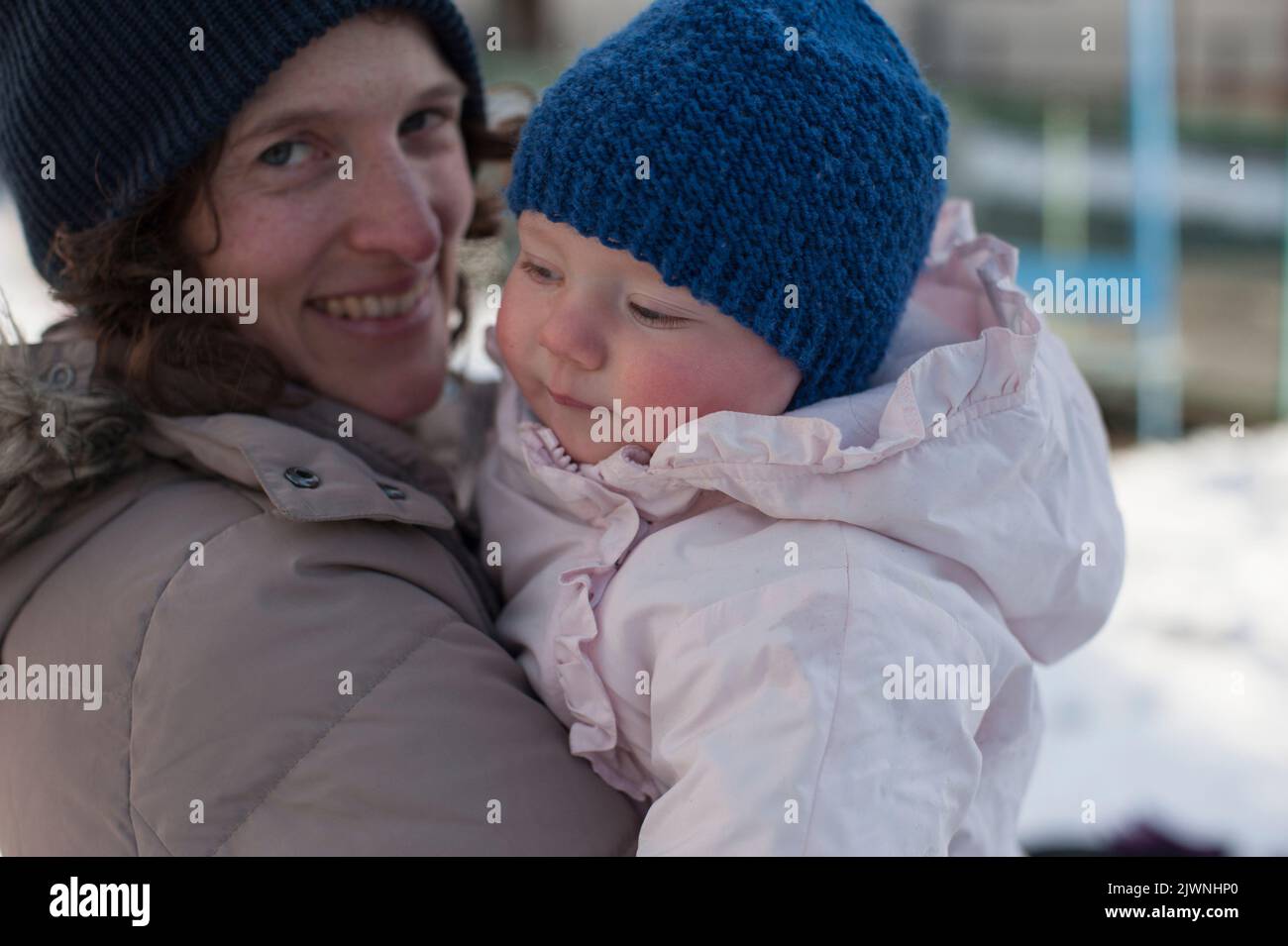 A smiling mother in winter gear holds a baby in a pink snowsuit and blue knitted touque hat against a wintry urban background. Stock Photo