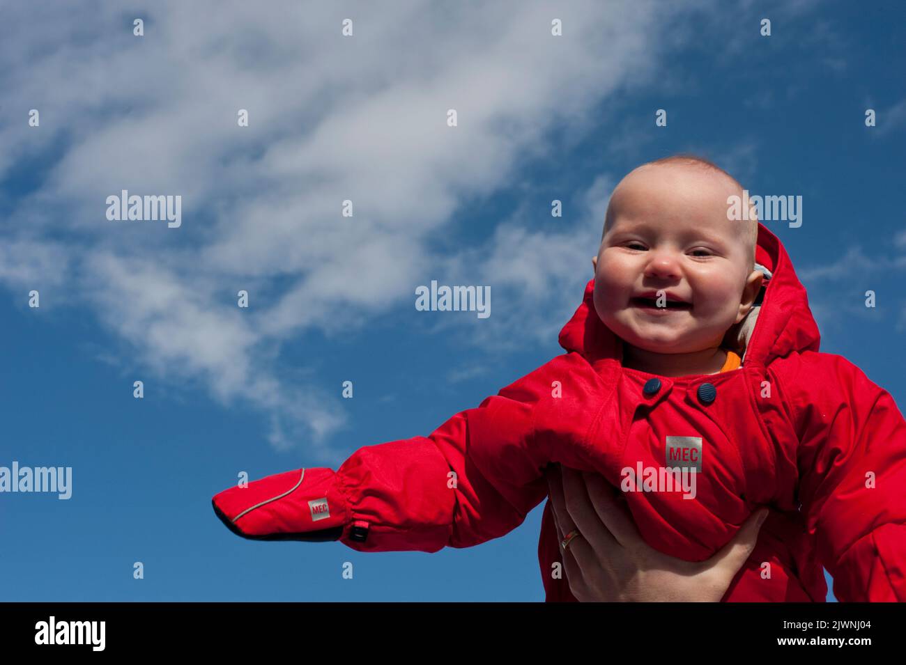 A nine-month-old baby in a red snowsuit and mittens is lifted up in her father’s hand againts the blue sky. Stock Photo