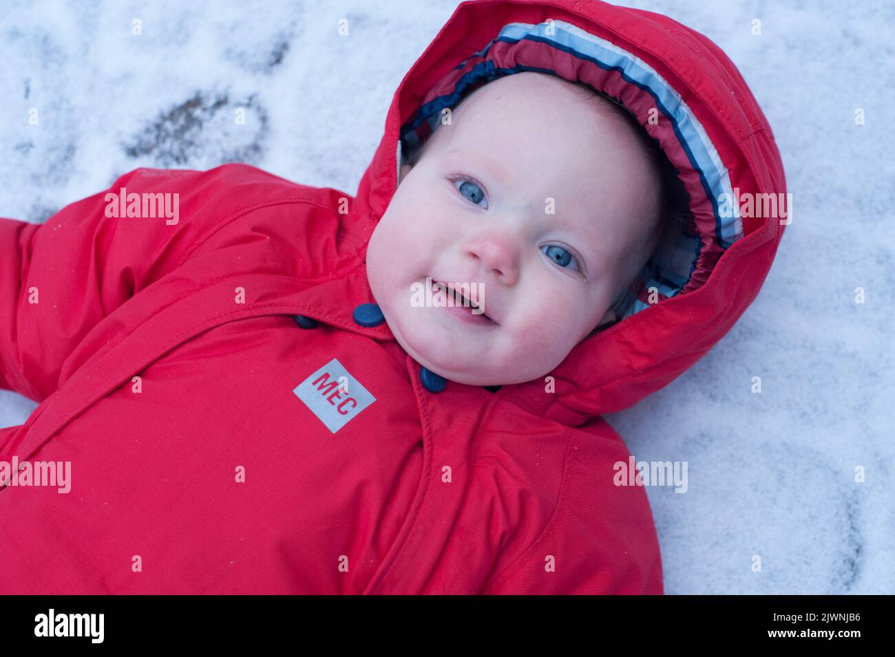 Portrait of a nine-month-old baby in a red snowsuit lying in the snow on a winter’s day. Stock Photo