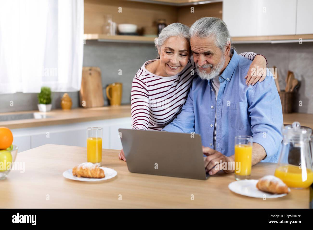 Happy elderly spouses spending time with laptop in kitchen Stock Photo