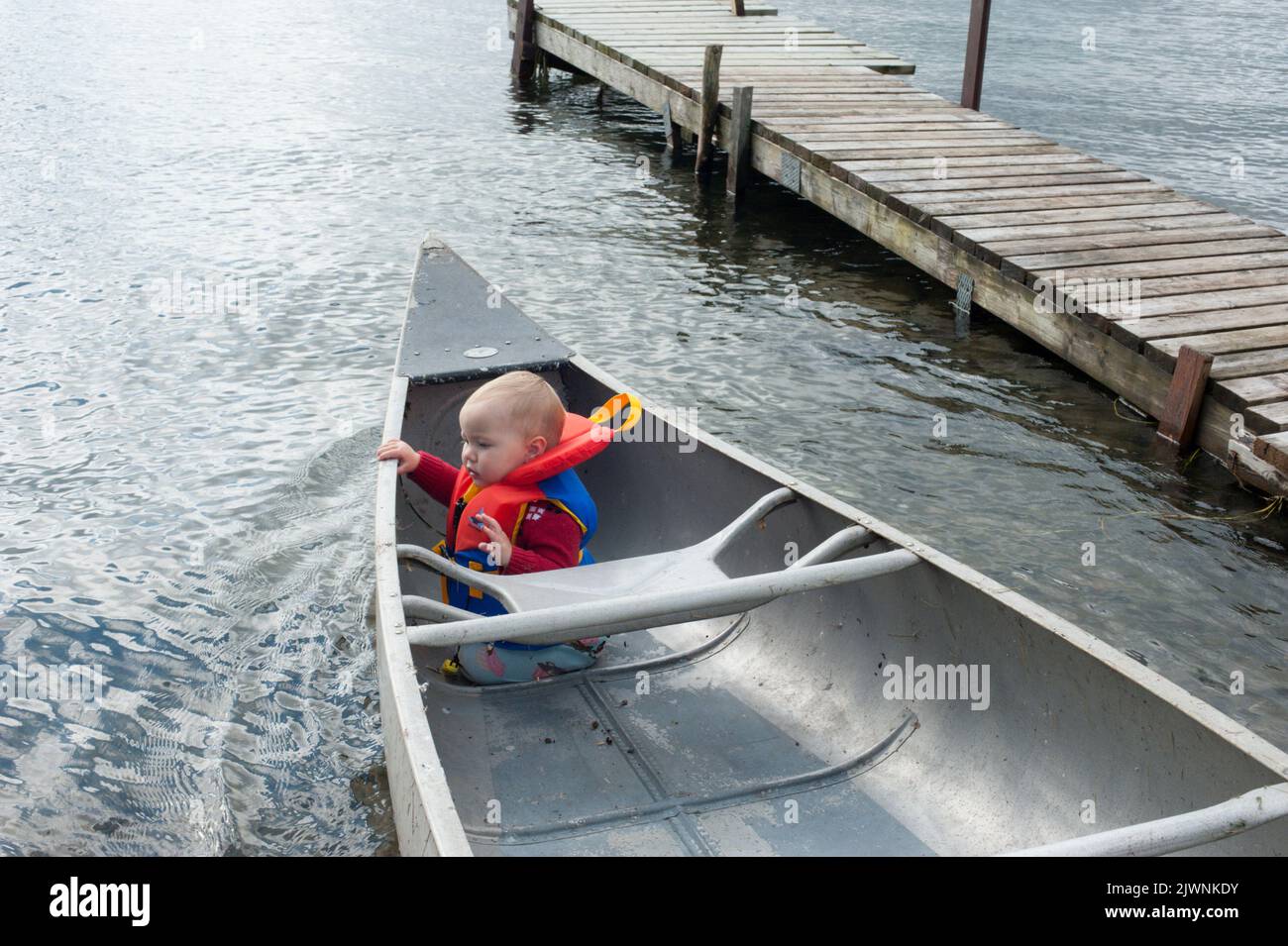 A one-year-old toddler sits in the bow of a grumman canoe by an old wooden dock on the Saint Lawernce River. Stock Photo