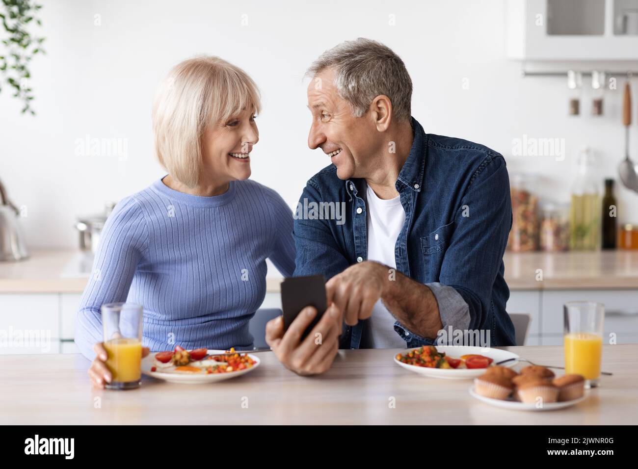 Smiling senior spouses having breakfast and using cell phone Stock Photo