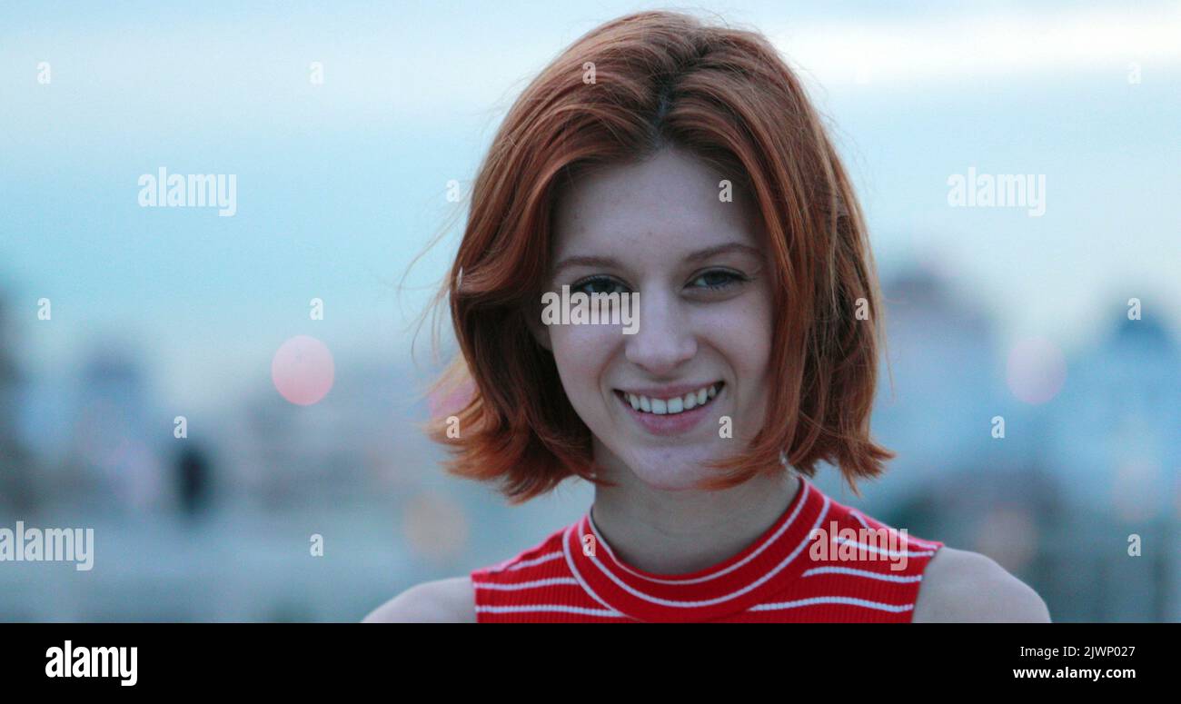 Portrait of young redhead millennial woman smiling to camera in city in background Stock Photo
