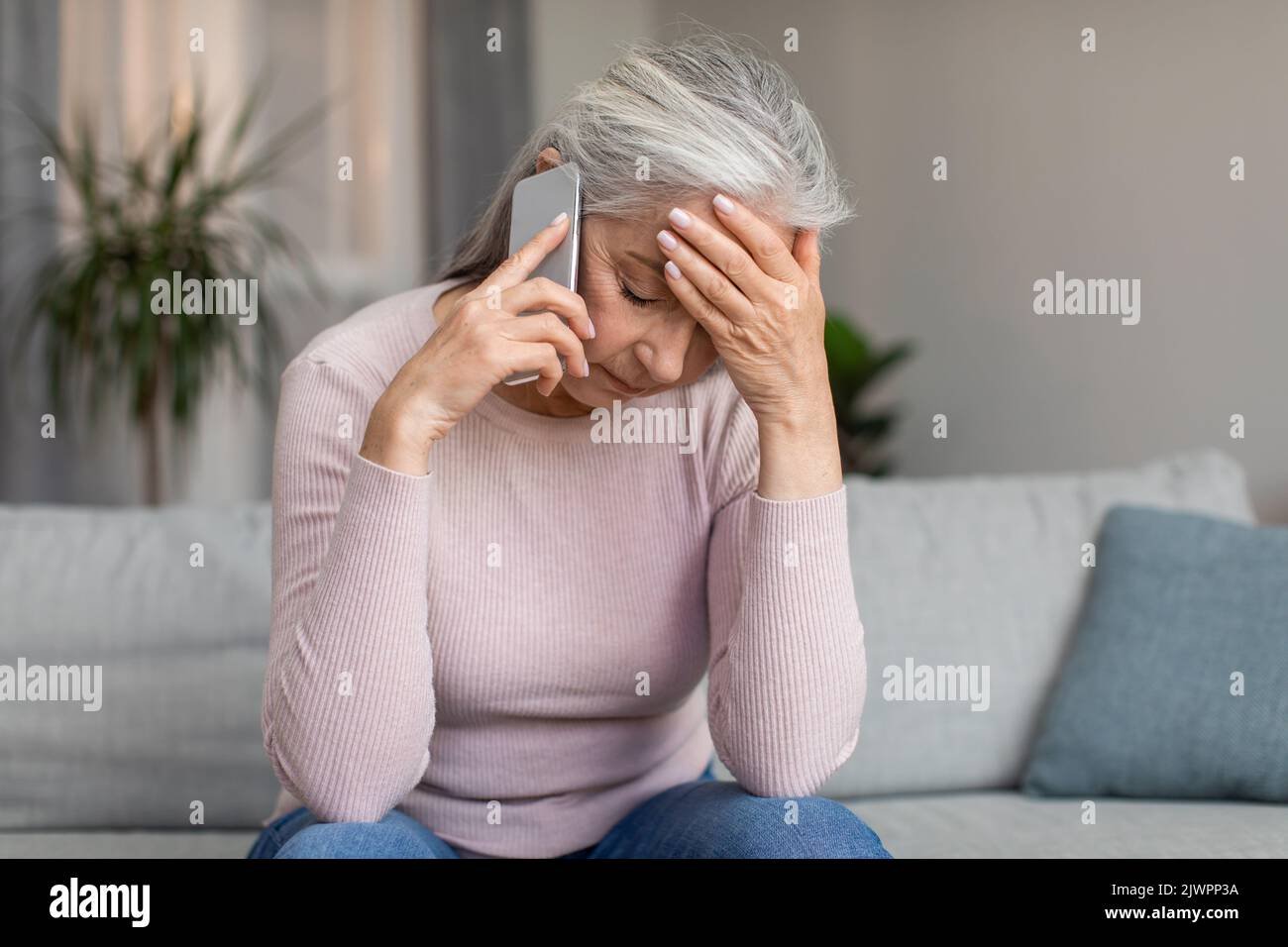 Depressed unhappy caucasian elderly gray-haired lady speaks by phone and holds her head with hand Stock Photo