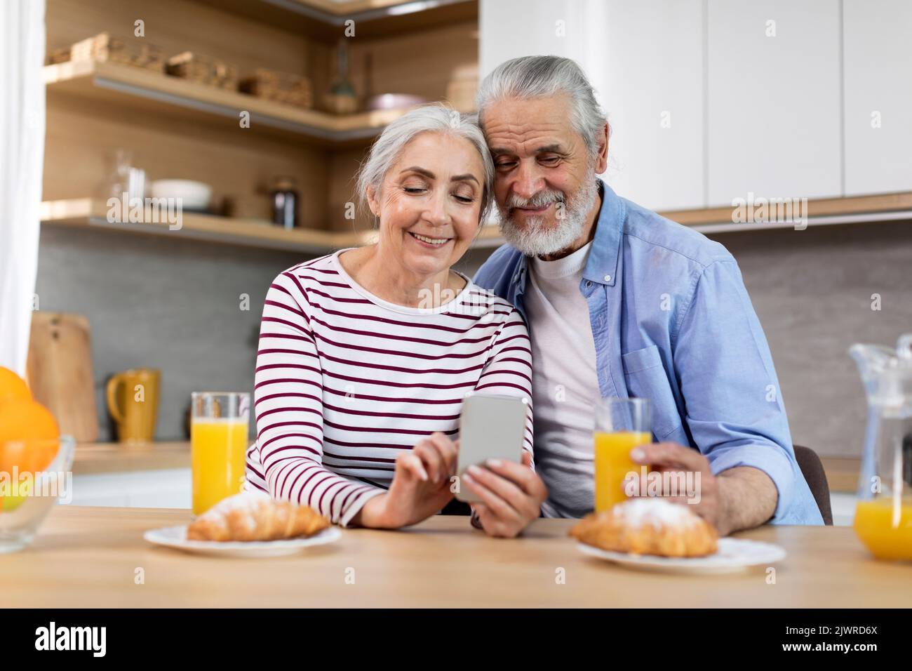 Happy Senior Spouses Spending Time With Smartphone While Having Breakfast In Kitchen Stock Photo
