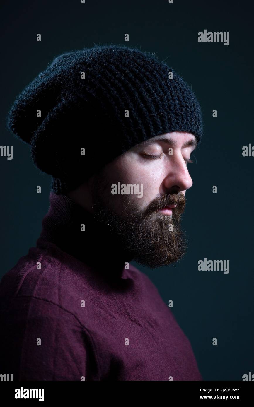 bearded guy with closed eyes in a knitted black hat and burgundy sweater. On a gray background. Stock Photo