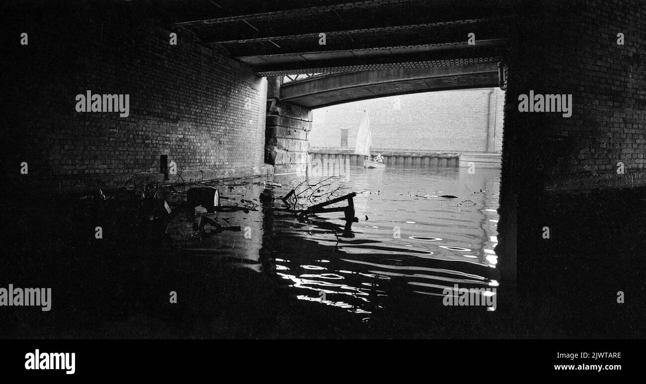London, England, circa 1967. A view from under a bridge over the Regent’s Canal. Two boys, who are members of The Pirate Club are sailing in a small dinghy in the distance. A large quantity of debris has collected under the bridge. The Pirate Club, a children’s boating club was formed in 1966 at Gilbey’s Wharf on the Regents Canal near Camden, London. Their clubhouse was an old barge and a number of small boats and canoes had been donated for the children’s use. Stock Photo