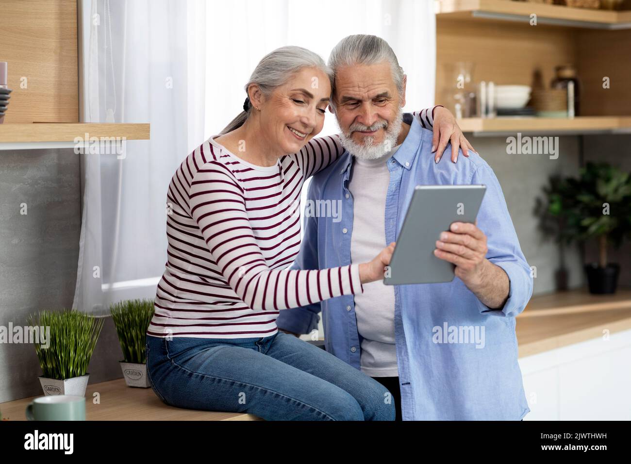 Happy Senior Spouses Using Digital Tablet While Relaxing In Kitchen Together Stock Photo