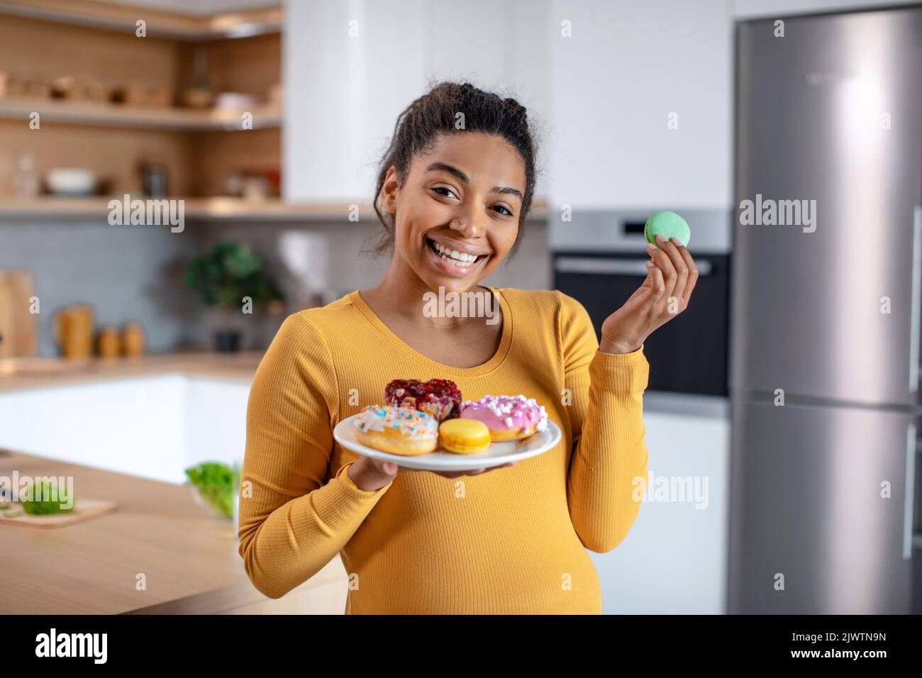 Cheerful young pregnant african american lady with belly hold plate of sweets in minimalist light kitchen interior Stock Photo