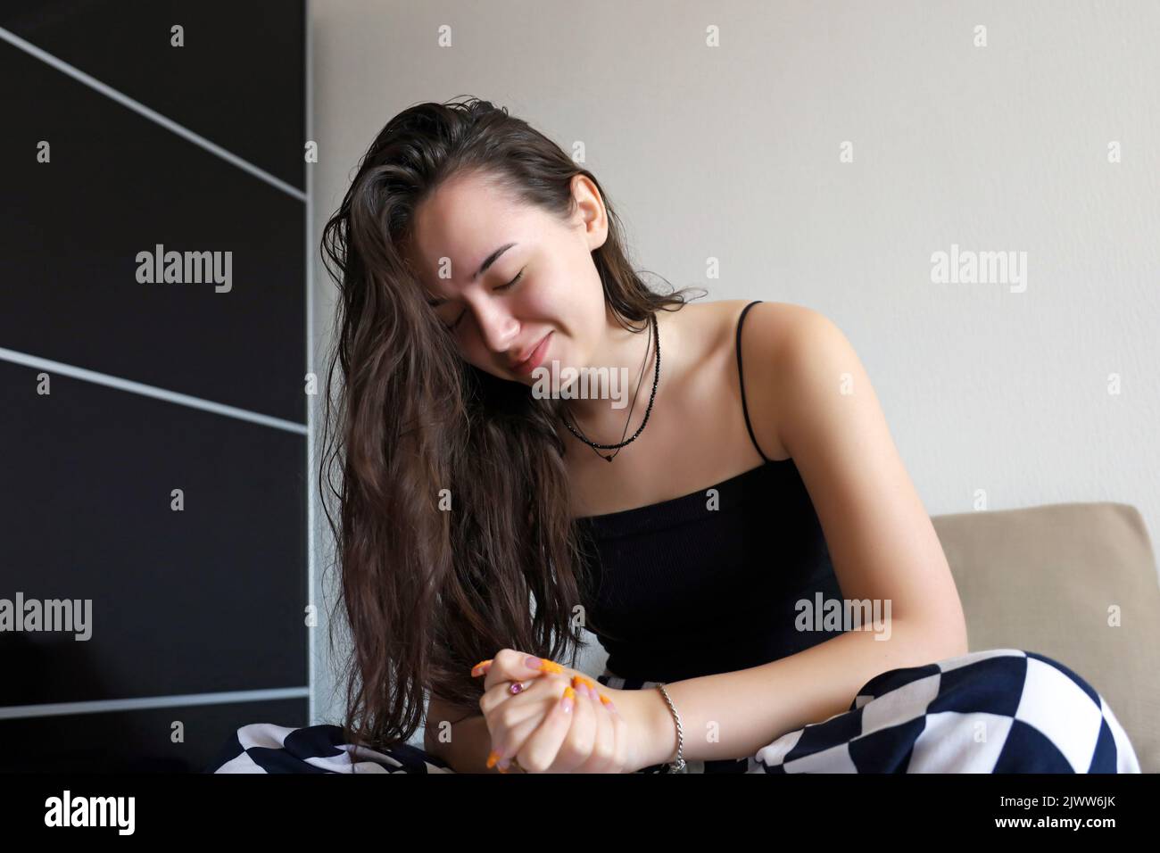 Pretty hopeful girl with long hair sitting with her eyes closed and hands clasped on sofa. Concept of prayer or making wishes Stock Photo