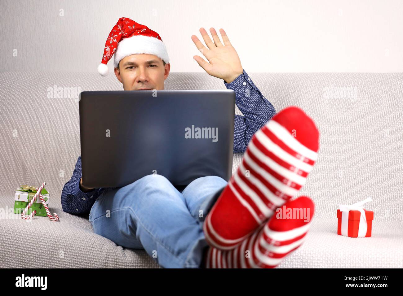 Man in Santa hat and Christmas socks sitting with laptop and gifts on a sofa with greeting gesture. Remote New Year celebration in home, online video Stock Photo
