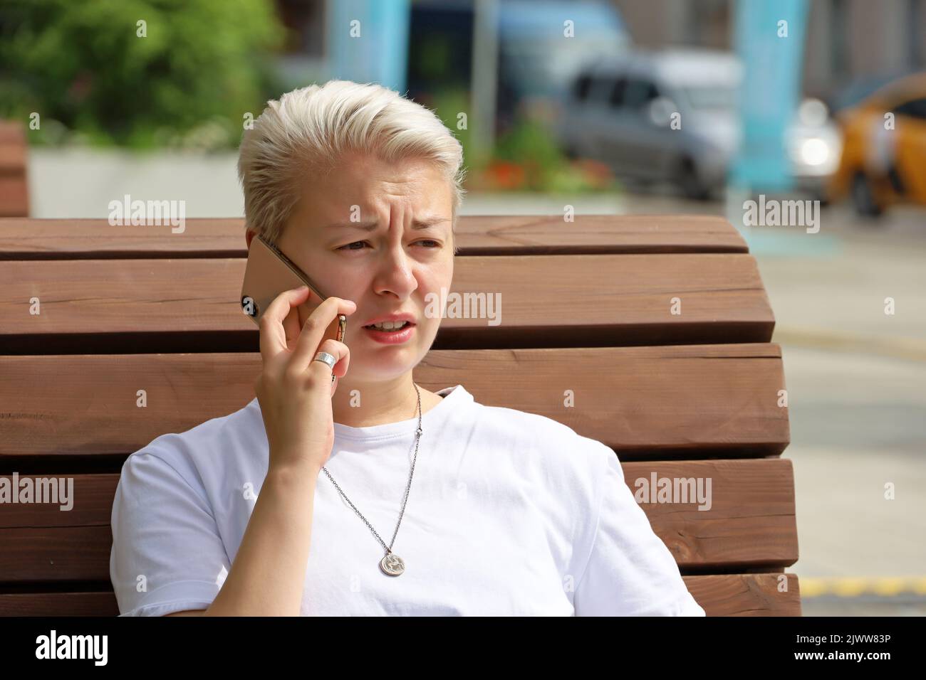 Girl with short blonde hair talking on mobile phone while sitting on street wooden bench. Tomboy lifestyle in summer or autumn city Stock Photo