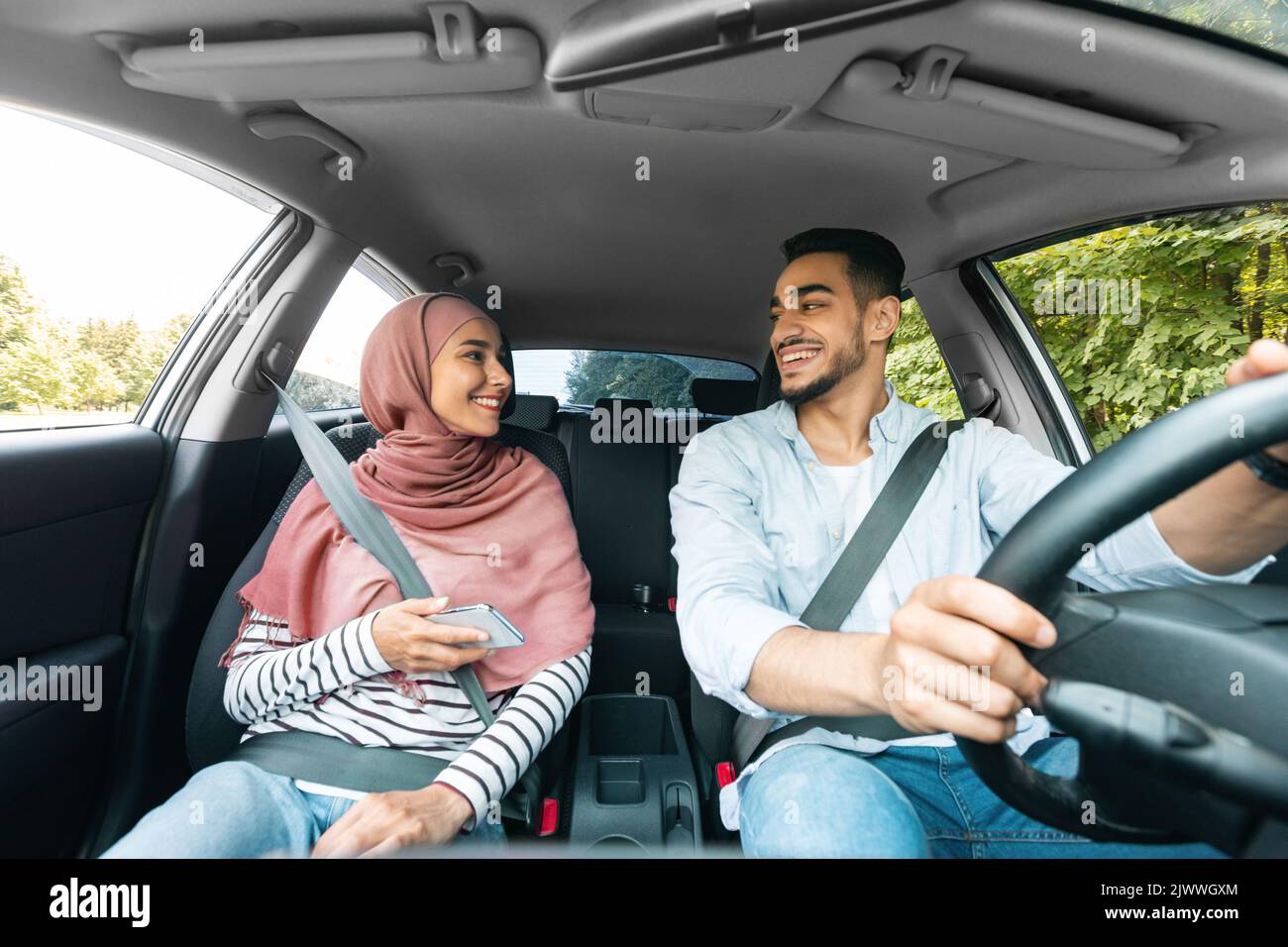 Cheerful young middle eastern woman in hijab with smartphone and husband rides on car Stock Photo